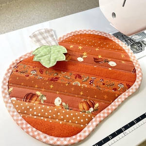 Puede incluir: Un posavasos acolchado con forma de calabaza con un tallo de cuadros vichy y una hoja verde. El posavasos est&aacute; hecho de tela naranja con una variedad de estampados oto&ntilde;ales, incluyendo calabazas, hojas y estrellas. El posavasos est&aacute; rodeado por un ribete de cuadros vichy blanco y rosa.