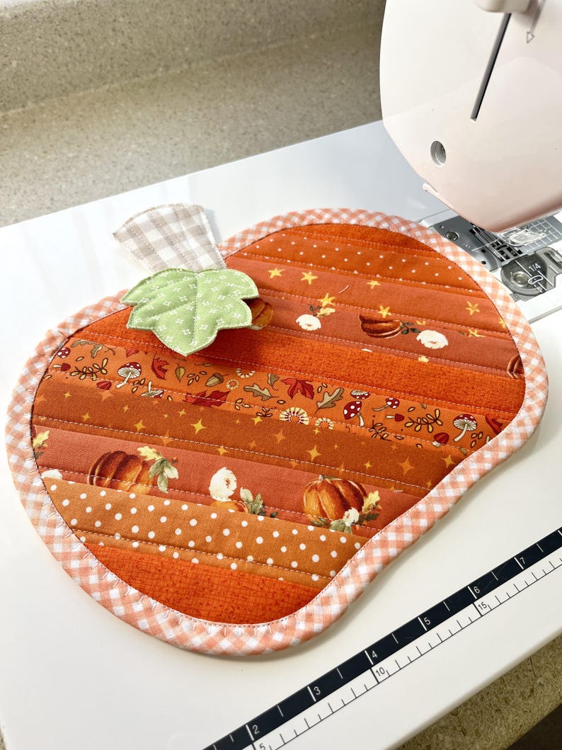 Puede incluir: Un posavasos acolchado con forma de calabaza con un tallo de cuadros vichy y una hoja verde. El posavasos est&aacute; hecho de tela naranja con una variedad de estampados oto&ntilde;ales, incluyendo calabazas, hojas y estrellas. El posavasos est&aacute; rodeado por un ribete de cuadros vichy blanco y rosa.