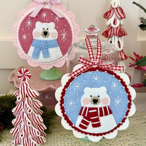 May include: Two circular felt ornaments featuring white polar bears. One bear wears a blue scarf on a pink background, the other a red and white striped scarf on a light blue background. Both have embroidered snowflakes, scalloped edges, and gingham ribbon bows.