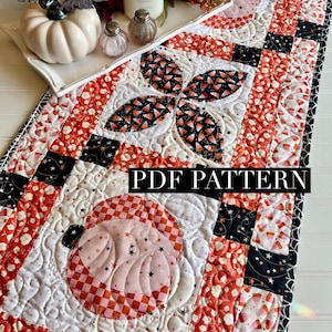以下が含まれることがあります： ハロウィーンをテーマにしたキルトテーブルランナー。 ランナーは、オレンジ、黒、白のパンプキンとキャンディーコーンのデザインが特徴です。 テキスト「PDF PATTERN」がランナーに表示されます。 ランナーの長さは約102cmです。
