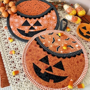 May include: Two round quilted fabric pot holders featuring jack-o'-lantern faces. The top holder has an orange spiderweb print with a black gingham border. The bottom holder has an orange floral pumpkin print with a white and orange gingham border.