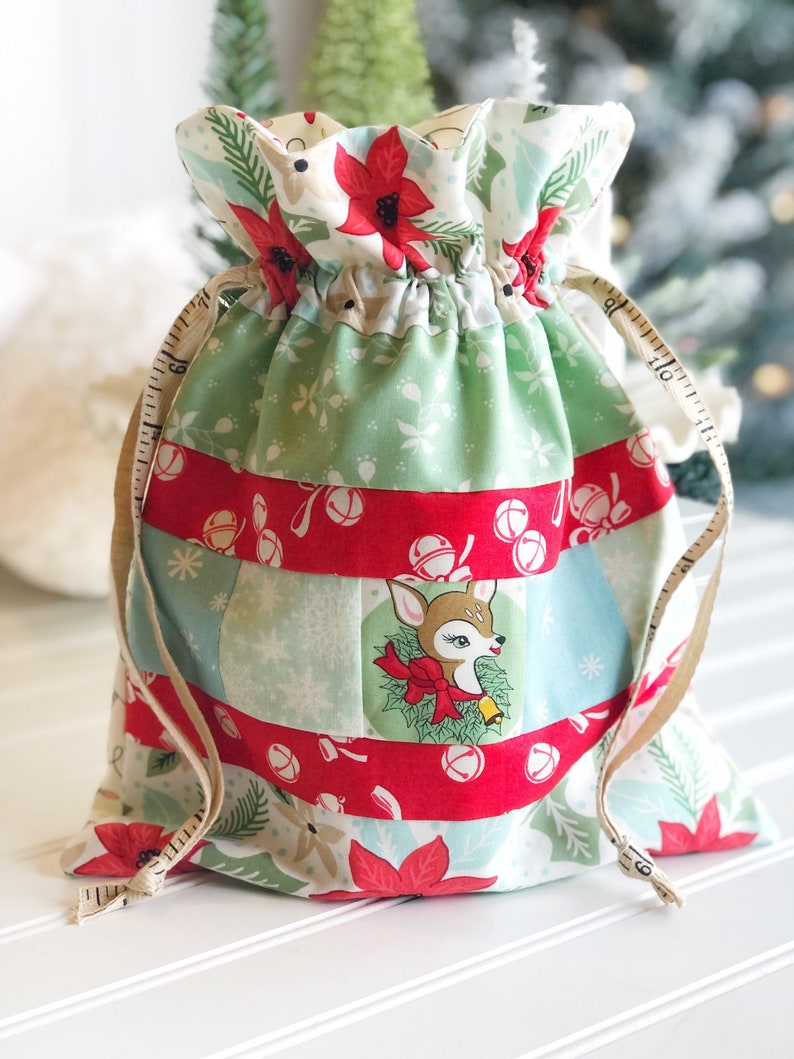 May include: A green and red Christmas gift bag with a deer and bells print. The bag has a drawstring closure and a white measuring tape tied to it.