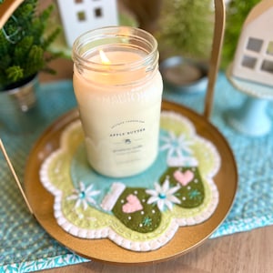 May include: A clear glass jar candle with a white label that reads "Brewton's Antique Candle Co. Apple Butter" and a lit wick. The candle is sitting on a green and white felt coaster with a snowflake design and two embroidered mittens with pink hearts.