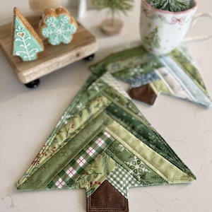 Puede incluir: Posavasos acolchados en forma de árbol de Navidad en varios tonos de verde, con un detalle de tronco marrón. Los posavasos están colocados sobre una superficie blanca, con una taza y árboles decorativos en el fondo. Los posavasos miden unos 15 cm de ancho.