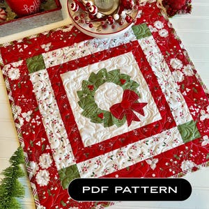 May include: A red and green quilted table runner with a wreath design in the centre. The runner has a white background with red and green floral patterns. The text "PDF PATTERN" is printed on the bottom of the runner.