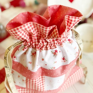 May include: A pink and white drawstring bag with a gingham pattern and floral print. The bag is made of fabric and has a white drawstring.