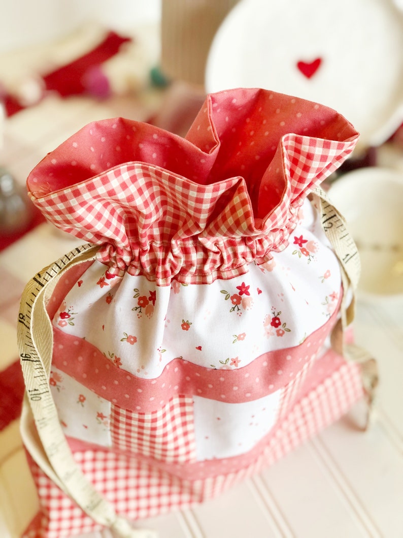 May include: A pink and white drawstring bag with a gingham pattern and floral print. The bag is made of fabric and has a white drawstring.
