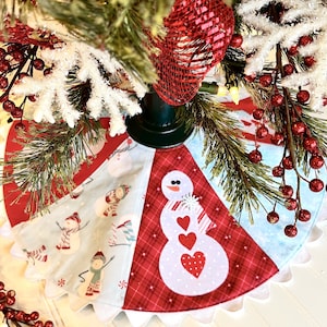 May include: A Christmas tree skirt with a scalloped edge. The skirt features a patchwork design with a snowman and snowflake pattern. The snowman has heart-shaped details. The skirt is red, white, and light blue.