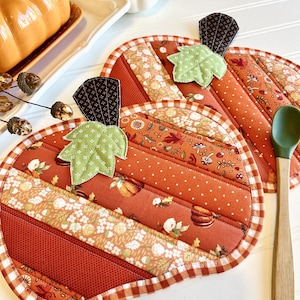 May include: Two quilted pumpkin-shaped pot holders with a gingham trim. The pot holders are made from patchwork fabric with a variety of fall-themed prints, including pumpkins, leaves, and acorns.