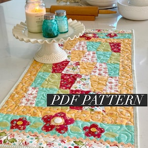 May include: A colorful quilted table runner with a patchwork design featuring floral patterns in red, yellow, and turquoise. The runner has a scalloped edge and the text "PDF PATTERN" is displayed on the front.