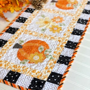 May include: A quilted table runner with orange pumpkin and yellow flower appliqués on a white background. The border is black and white check, with orange and black striped trim. The runner is ideal for autumn or fall home decor.