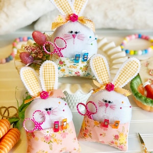 May include: Three white felt bunny pincushions with yellow and white gingham ears, pink floral fabric pockets, and pink and blue accents. Each bunny has a pair of pink scissors, safety pins, and binder clips in its pocket.