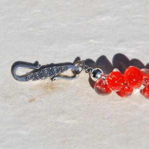 May include: A silver clasp with a decorative swirl design is attached to a strand of red and clear glass beads.