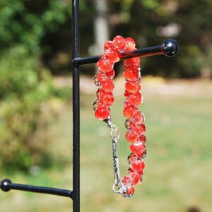 May include: A red glass bead bracelet with a silver clasp. The bracelet is hanging on a black metal stand.