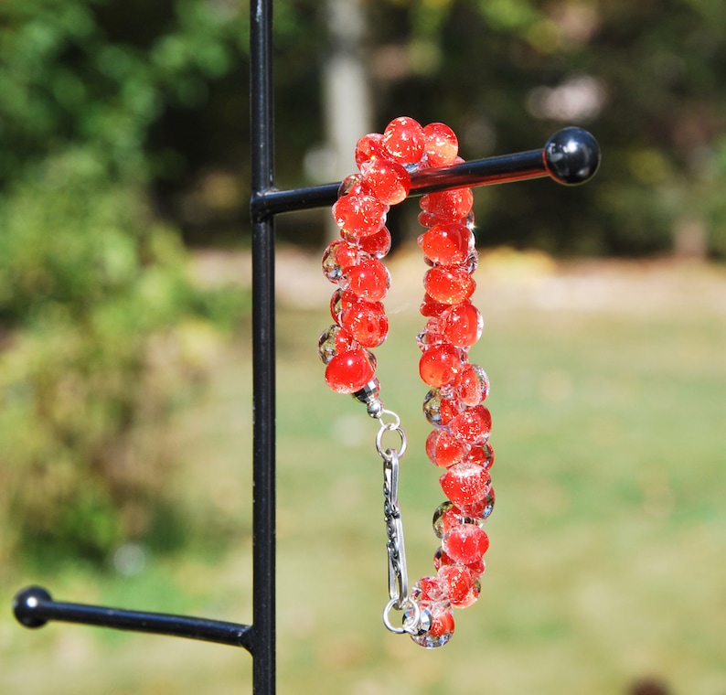 May include: A red glass bead bracelet with a silver clasp. The bracelet is hanging on a black metal stand.