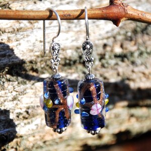 May include: A pair of handcrafted earrings with dark blue glass beads. The beads have a brown, swirling design and are accented with small, colorful glass beads. The earrings have silver-colored metal hooks and decorative tops, creating a unique and artistic accessory.