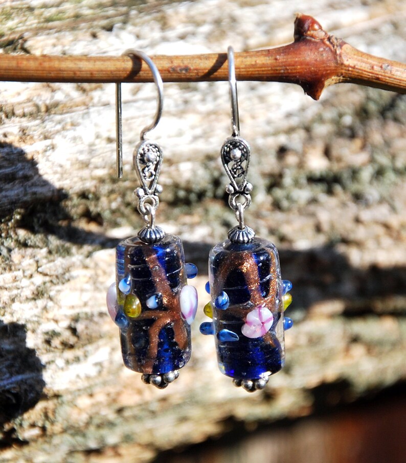 May include: A pair of handcrafted earrings with dark blue glass beads. The beads have a brown, swirling design and are accented with small, colorful glass beads. The earrings have silver-colored metal hooks and decorative tops, creating a unique and artistic accessory.