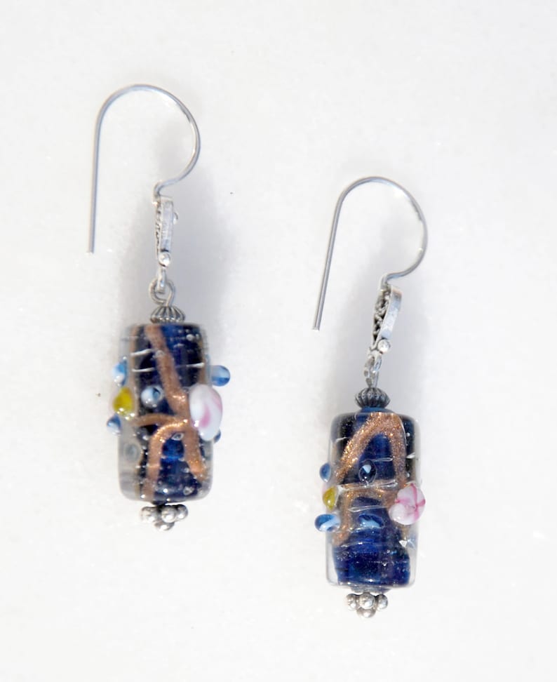 May include: A pair of dangle earrings featuring dark blue, cylindrical glass beads with copper-colored swirls and small, colorful accents. The earrings have silver-toned ear wires and decorative bead caps.
