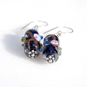 May include: A pair of handcrafted glass earrings. Each earring features a dark blue, oval-shaped bead with brown and gold accents, small pink and blue details, and a cluster of silver-colored beads at the base. The earrings have silver-colored ear wires.