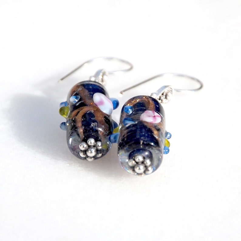 May include: A pair of handcrafted glass earrings. Each earring features a dark blue, oval-shaped bead with brown and gold accents, small pink and blue details, and a cluster of silver-colored beads at the base. The earrings have silver-colored ear wires.