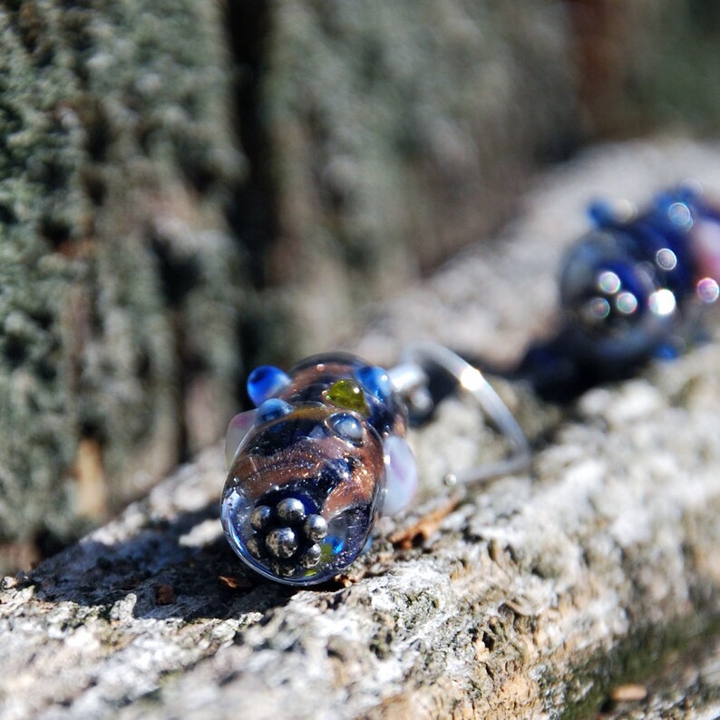 May include: Close-up of a pair of artisan glass earrings. The earrings feature a combination of clear, blue, brown, and green glass elements. The design includes small, silver-colored beads and a unique, organic shape. The earrings are displayed on a textured, wooden surface.