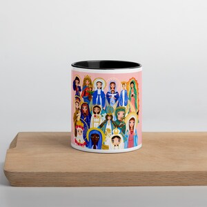 May include: A white ceramic mug with a black rim and handle. The mug features a colorful illustration of various depictions of the Virgin Mary. The background is a light pink color.