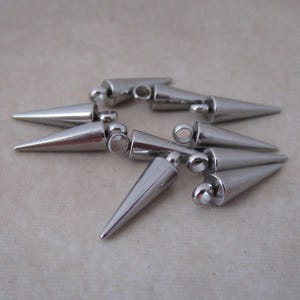 May include: A collection of silver-colored metal cone-shaped charms with a pointed end and a small loop at the top. The charms are arranged on a neutral-toned surface, reflecting light.