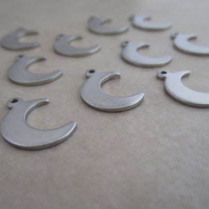 May include: A group of silver crescent moon charms, scattered on a light brown surface. The charms are all the same size and shape, and they have a smooth, polished finish.