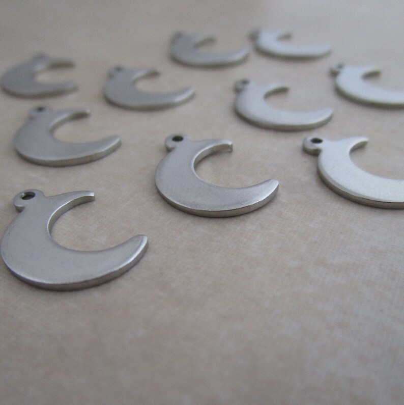 May include: A group of silver crescent moon charms, scattered on a light brown surface. The charms are all the same size and shape, and they have a smooth, polished finish.
