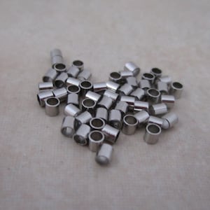 May include: A collection of small, cylindrical silver-colored metal beads. Each bead has a hole running through the center. The beads are scattered on a light beige surface.