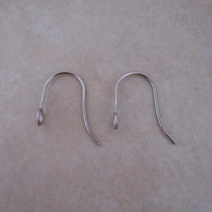 May include: Two silver-colored earring hooks. Each hook has a curved top and a straight, slightly angled bottom. The hooks have a small loop at the top and are designed for pierced ears. The hooks are made of a shiny metal.