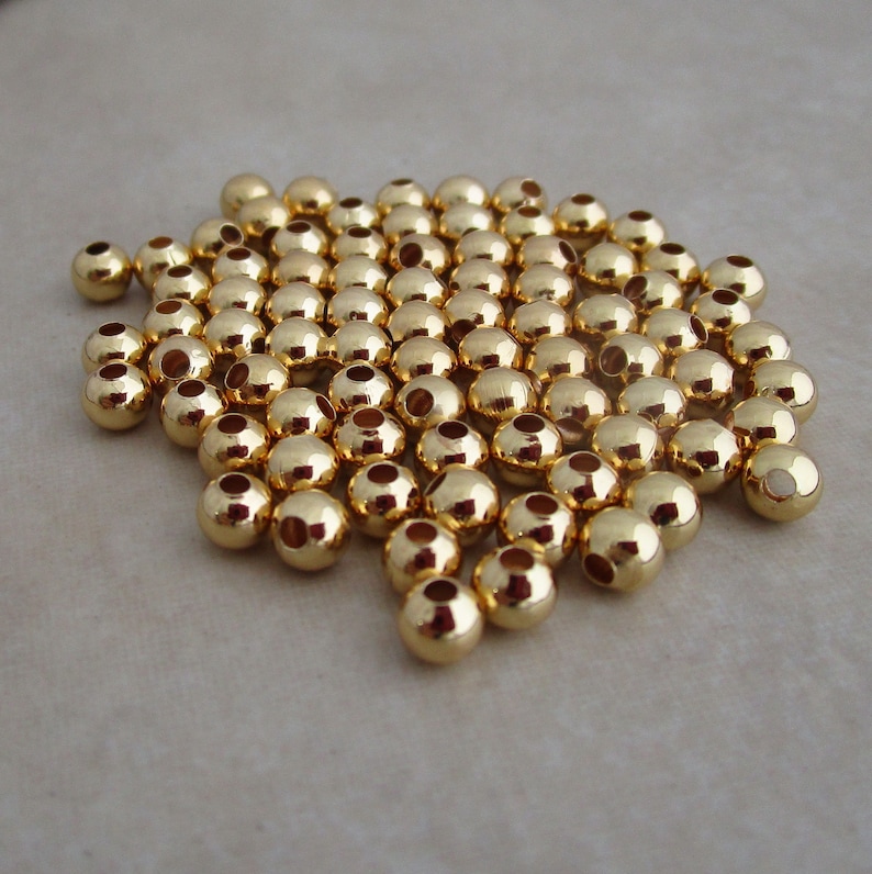 May include: A pile of small, shiny, gold-colored round beads. Each bead has a hole through the center. The beads are scattered on a light-colored surface, reflecting light.