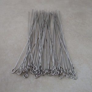 May include: A collection of silver-colored head pins with looped ends. The pins are thin and straight, lying in a scattered arrangement against a neutral background. These pins are likely used for jewelry making or crafting.
