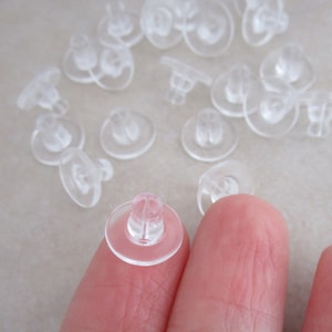 May include: A close-up of a clear plastic earring back. The earring back has a small post that fits into the earring and a larger disc that sits on the back of the ear.