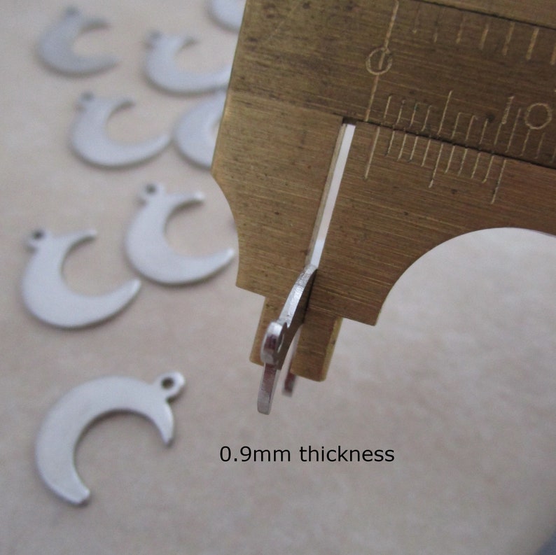May include: Close-up of a silver crescent moon charm measured with calipers. The charm is 0.9mm thick.