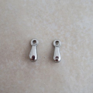May include: Two polished silver teardrop-shaped charms with a small loop at the top. The charms are smooth and reflective, ideal for jewelry making. They are displayed on a light beige surface.