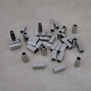 May include: A collection of small, rectangular metal tubes with a hollow center. The tubes are silver in color and appear to be made of stainless steel. They are scattered across a light gray surface.