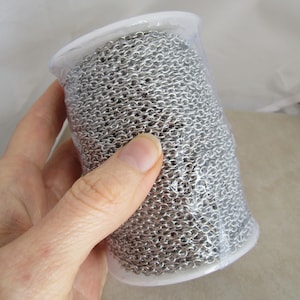 May include: A roll of silver chain in a clear plastic container. The chain is made of small, linked metal pieces.