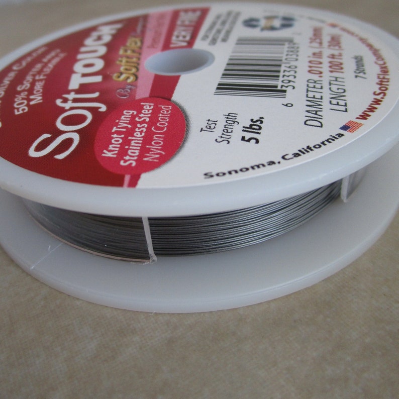 May include: A spool of silver-colored Soft Touch knot tying wire. The label reads "Soft Touch" and "Knot Tying by Soft Flex" with details about the wire: "Stainless Steel, Nylon Coated", "Test Strength 5 lbs", "Diameter .010 in. (2.5mm)", "Length 100 ft. (30m)", "7 Strands", "Sonoma, California", and "www.SoftFlexCompany.com".