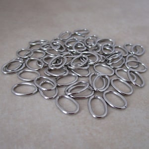 May include: A pile of small, shiny, silver-colored oval rings. These metal rings are smooth and have a consistent shape, suitable for various crafting or jewelry-making projects. The rings are scattered on a neutral-toned surface.