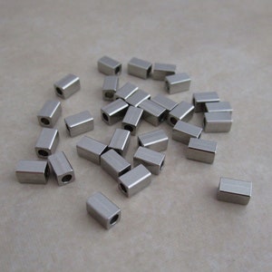 May include: A group of silver metal spacer beads, each with a rectangular shape and a hole in the center. The beads are scattered on a light brown surface.