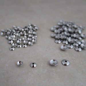 May include: A close-up of two piles of silver metal bead caps, with four individual caps in the foreground. The caps are round with a flat top and a slightly raised edge.