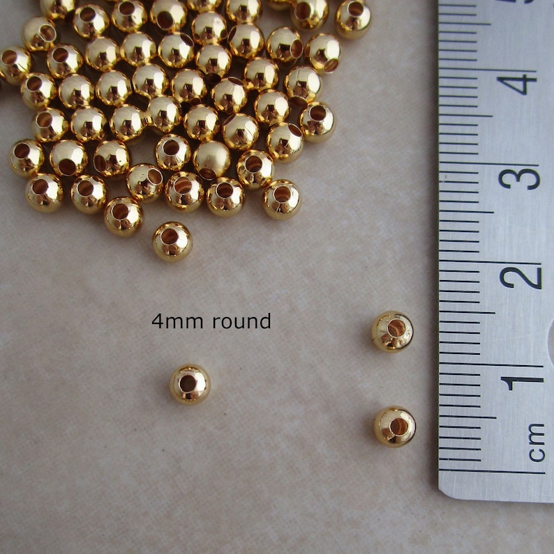 May include: A collection of shiny, gold-colored, round beads, each approximately 0.16 inches in diameter, are scattered across a light-colored surface. The text "4mm round" is visible. A metal ruler with centimeter markings is in the frame.