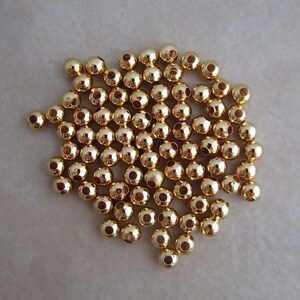 May include: A collection of small, shiny, gold-colored beads. Each bead is spherical with a hole running through the center. The beads are scattered on a neutral-toned surface, reflecting light and creating a bright, metallic appearance. Ideal for jewelry making and crafts.