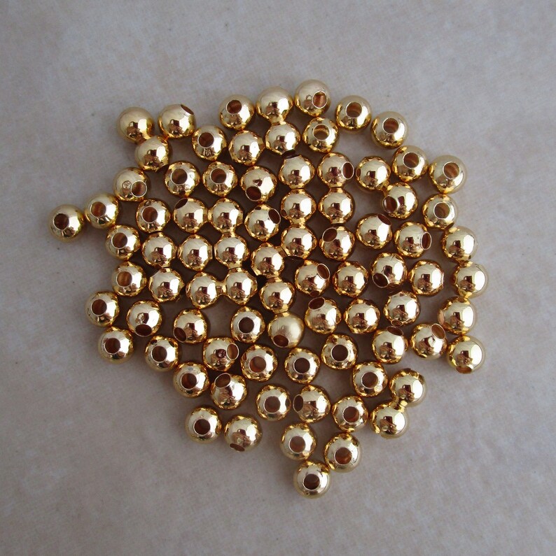 May include: A collection of small, shiny, gold-colored beads. Each bead is spherical with a hole running through the center. The beads are scattered on a neutral-toned surface, reflecting light and creating a bright, metallic appearance. Ideal for jewelry making and crafts.