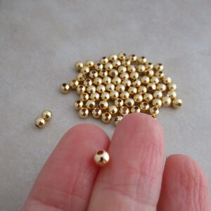 May include: A collection of small, shiny, gold-colored beads. The beads are spherical with a small hole through the center. Some beads are scattered, while others are held between fingers. These beads are ideal for jewelry making and crafting.