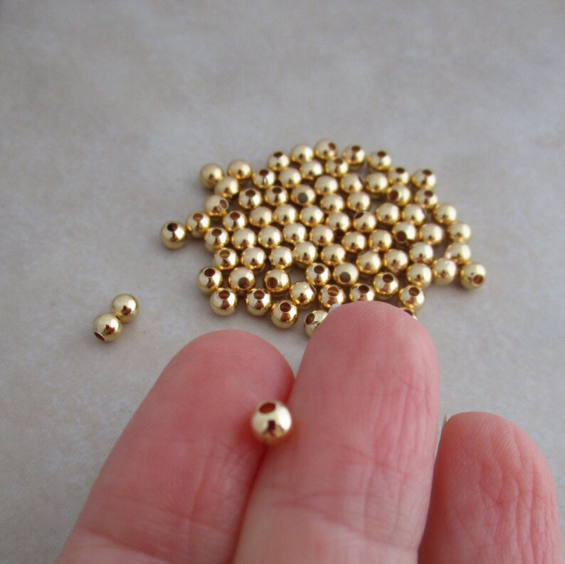 May include: A collection of small, shiny, gold-colored beads. The beads are spherical with a small hole through the center. Some beads are scattered, while others are held between fingers. These beads are ideal for jewelry making and crafting.