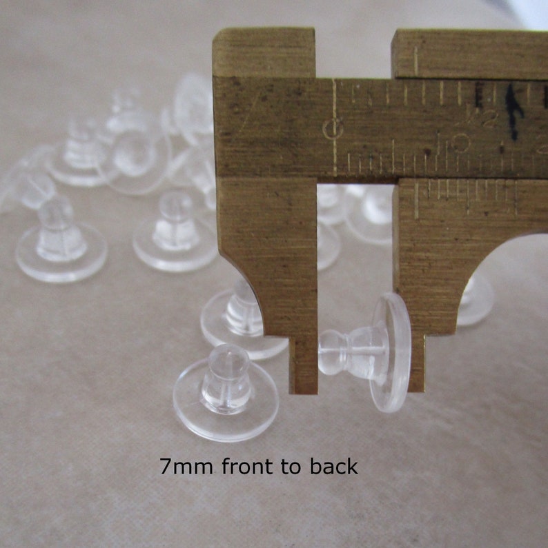 May include: Close-up of a caliper measuring the height of a clear plastic earring back. The caliper reads 7mm front to back.