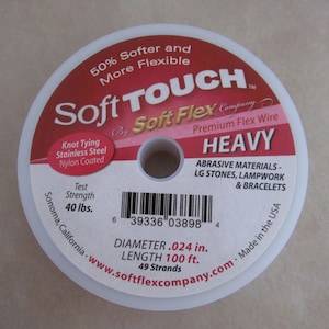 May include: A spool of Soft Touch by Soft Flex Company Heavy Premium Flex Wire. The spool is white with red text and a barcode. The text on the spool reads "50% Softer and More Flexible", "Knot Tying Stainless Steel Nylon Coated", "Test Strength 40 lbs.", "Abrasive Materials - LG Stones, Lampwork & Bracelets", "Diameter .024 in.", "Length 100 ft.", "49 Strands", "Made in the USA", and "www.softflexcompany.com".