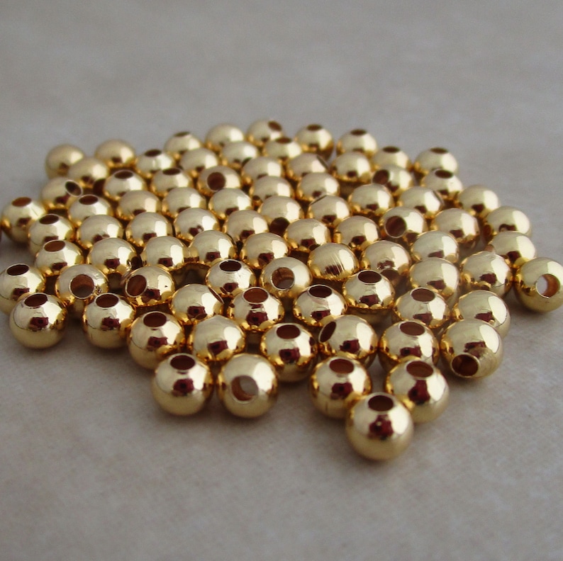 May include: A collection of small, shiny, gold-colored beads. Each bead is round with a hole through the center, reflecting light. The beads are scattered, suggesting they are loose and ready for use in jewelry or crafts.